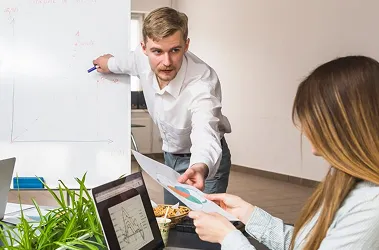 A man and woman collaborate on a whiteboard, discussing ideas and writing notes in a professional setting.